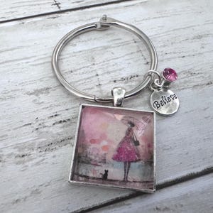 May include: A silver-toned metal keychain featuring a square pendant with a printed illustration of a person in a pink dress holding an umbrella with a small cat nearby. The keychain includes a small round charm engraved with the word Believe and a pink crystal.