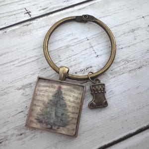 May include: A bronze-toned metal keychain featuring a circular ring, a square pendant with a printed image of a Christmas tree over musical notes, and a small dangling charm shaped like a Christmas stocking with a decorative cuff detail.