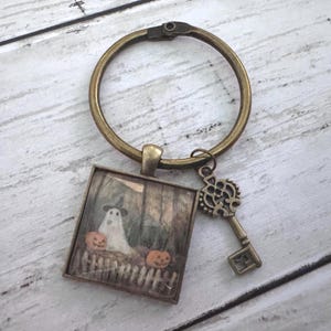 May include: A bronze-toned metal keyring featuring a square pendant with a printed image of a ghost wearing a hat standing behind a fence with two orange pumpkins. An ornate, skeleton-style key charm hangs from the same circular ring.