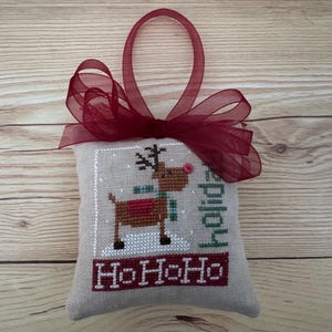 May include: A cross-stitched Christmas ornament featuring a reindeer with a red nose and scarf, standing on snow. The ornament has the word "holiday" and "HoHoHo" stitched on it. A red ribbon is attached for hanging.