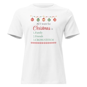 Può includere: T-shirt bianca a tema natalizio. La maglietta presenta una ghirlanda di ornamenti e il testo "All I want for Christmas is" seguito da "Family, Friends, CROSS STITCH."