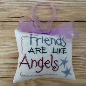 May include: A small, white, cross-stitched pillow with the words "Friends ARE LIKE Angels" in black and burgundy thread. Two blue stars are stitched on the right side. A lavender ribbon is tied at the top for hanging.
