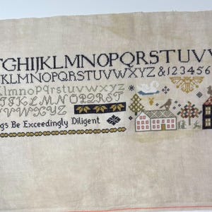 May include: Cross-stitch sampler on beige fabric, featuring the alphabet, numbers, and decorative motifs. The design includes houses, trees, birds, and the text "In All Things Be Exceedingly Diligent". The letters and numbers are in black and gray.
