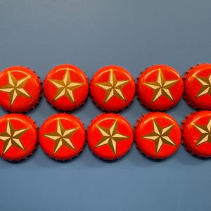 May include: Ten red bottle caps with a white and gold star design arranged in two rows on a blue surface. The caps are evenly spaced, showcasing the star motif.