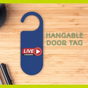 May include: A blue hangable door tag with the words "LIVE STREAMING" and a play button icon. The tag also has the words "HANGABLE DOOR TAG" in green. The tag is on a wooden surface.