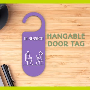 Può includere: Un cartello da porta viola con la scritta "IN SESSION" e un'immagine di due figure stilizzate sedute su sedie. Le parole "HANGABLE DOOR TAG" sono in verde. Il cartello è su una superficie di legno.