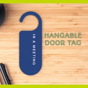 May include: A blue door tag with the text "IN A MEETING" and "HANGABLE DOOR TAG" in green. The tag is on a wooden surface, with a pen, notepad, and a cup of coffee in the background.