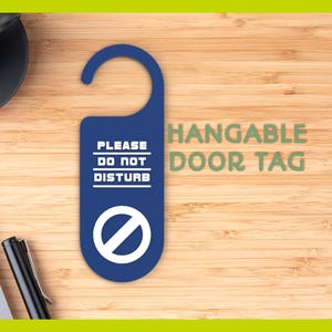 Könnte beinhalten: Ein blauer Türanhänger mit der Aufschrift "PLEASE DO NOT DISTURB" und einem weißen Symbol. Die Worte "HANGABLE DOOR TAG" sind grün. Der Anhänger liegt auf einer Holzoberfläche, mit einer Kaffeetasse und einem Stift.