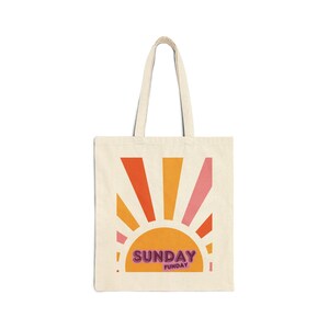 May include: A beige tote bag with a graphic of a yellow sun with orange and pink rays. The text "SUNDAY FUNDAY" is printed in pink and purple letters on the sun.