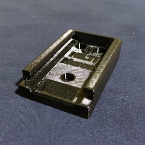 Puede incluir: Un soporte de plástico rectangular negro con el texto "HILTI" en relieve en la superficie. El soporte tiene un área empotrada con un agujero circular y bordes elevados. El artículo parece estar impreso en 3D.