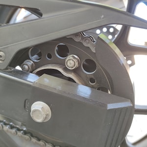 Op de afbeelding: Close-up van een motorachterwiel met een zwarte kettingbeschermer en een zilveren ketting. De kettingbeschermer heeft een getextureerd oppervlak en een verhoogde lip.