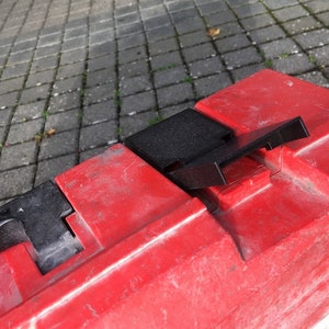 Puede incluir: Un contenedor de almacenamiento de plástico rojo con una tapa negra. La tapa tiene una pequeña manija negra en la parte superior.