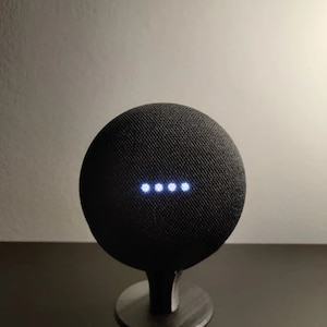 May include: A black Google Home smart speaker with a fabric covering and a white light ring around the top. The speaker is on a black stand.