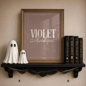 Pode incluir: Uma moldura retangular com um inserto malva apresentando o texto branco VIOLET Mockups, duas figuras de fantasmas de cerâmica branca e quatro livros pretos com texto branco em uma prateleira preta decorada com teias e aranhas.