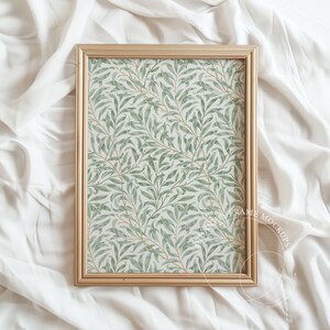 May include: A gold-coloured rectangular picture frame displaying a botanical print of green leaves and branches on a white background. The frame rests on white, textured fabric.