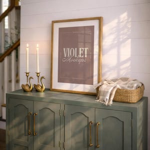 May include: A framed vertical poster with a mauve background and the white text VIOLET Mockups, displayed on a sage green cabinet with two brass swan candle holders and a woven basket containing a beige and white plaid throw blanket.