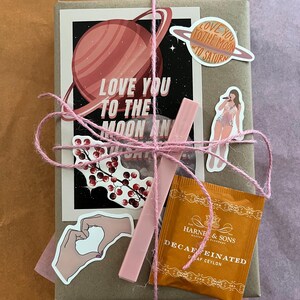 May include: A brown paper package wrapped with twine and decorated with stickers. The stickers feature a pink planet with rings, a pink heart, a pink floral design, a pink sticker with the text "Love you to the moon and to Saturn", and a pink sticker with a woman singing. The package also includes a pink stick and a small orange tea bag with the text "Harney & Sons" and "Decaffeinated" on it.