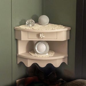 May include: A corner shelf unit painted a light beige color, featuring two shelves and a small drawer. The shelves are adorned with decorative doilies, and the top shelf holds a gray sphere and a clear crystal pumpkin. A gray speaker sits on the lower shelf.