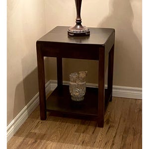 Peut inclure: Une table d'appoint carrée en bois brun foncé avec quatre pieds droits et une étagère inférieure. Un pied de lampe brun foncé repose sur le dessus, et un vase en verre transparent rempli de pierres décoratives et d'une fleur claire est posé sur l'étagère.