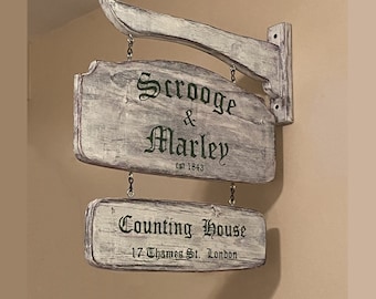 May include: A whitewashed wooden sign with black lettering that reads "Scrooge & Marley est 1843 Counting House 17 Thames St. London". The sign is hanging from a wooden arm.