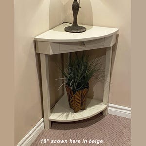 May include: Beige corner table with a drawer and a lower shelf. The table has a semi-circular design and is 45.7 cm wide. A decorative lamp sits on top, and a potted plant is on the lower shelf. The text "18" shown here in beige" is at the bottom.