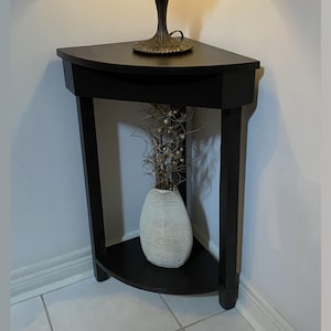 Peut inclure: Une table d'angle noire avec une étagère semi-circulaire et une étagère supérieure. Un vase blanc texturé avec des branches décoratives est posé sur l'étagère inférieure. Une lampe avec une base en bronze est posée sur l'étagère supérieure.