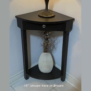 Peut inclure: Une table d'angle marron foncé avec un plateau triangulaire, un tiroir unique doté d'un bouton argenté et une étagère inférieure. La table mesure 38,1 cm. Un vase blanc avec des brindilles décoratives repose sur l'étagère inférieure.