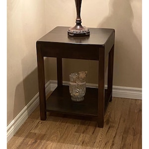May include: A square dark brown wooden end table featuring four straight legs and a lower shelf. The table has a smooth top surface and a clean, simple frame design. A glass vase filled with decorative stones sits on the lower shelf of the table.