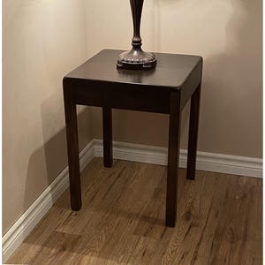May include: A square side table with a dark brown wood finish and four straight legs. The table features a smooth, flat top surface with rounded corners. A dark-coloured lamp base is positioned in the centre of the table top surface.