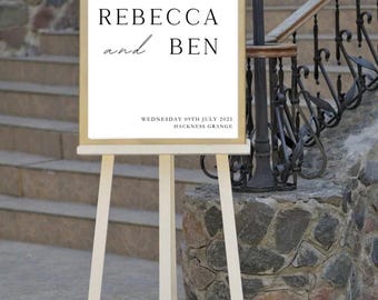 May include: A rectangular wedding welcome sign with a gold frame, displayed on a light-coloured wooden easel. The white sign features black text reading: WELCOME TO THE WEDDING OF REBECCA and BEN WEDNESDAY 09TH JULY 2025 HACKNESS GRANGE in a serif font.