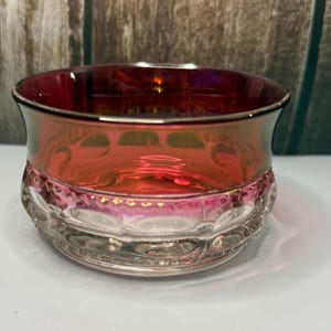 May include: A round glass bowl featuring a deep red iridescent finish on the upper portion and a clear, textured base with a repeating pattern of circular indentations and a horizontal band of small, raised geometric shapes around the middle.