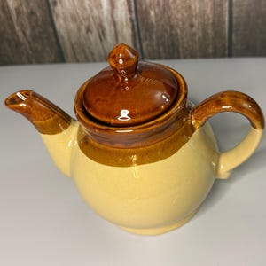May include: A ceramic teapot featuring a two-tone design with a glossy dark brown glaze on the lid, upper rim, spout, and handle, contrasted against a smooth, matte light beige body. The teapot includes a removable lid with a small knob.