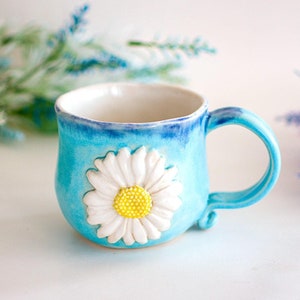 May include: A blue ceramic mug with a white daisy flower design. The mug has a handle and a rounded shape.
