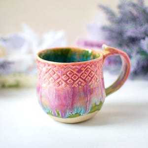 Puede incluir: Una taza de cerámica hecha a mano con un patrón de diamantes texturizado. La taza presenta una vibrante paleta de colores rosa, verde y azul. El asa tiene forma curva y el interior es de color verde azulado oscuro. Una pieza única de arte funcional.