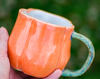 May include: An orange ceramic mug with a textured surface and a white interior. The mug has a rounded shape with a blue and green handle. The mug is held in a hand against a blurred green background.