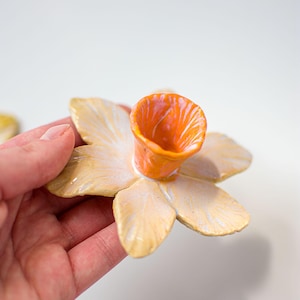 May include: A ceramic daffodil-shaped candle holder. The petals are a creamy yellow with iridescent accents, and the center is a vibrant orange. The candle holder is designed to resemble a flower.
