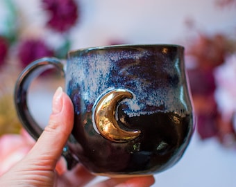 May include: A ceramic mug with a rounded body and a curved handle. The exterior features a dark, mottled glaze in shades of deep blue, purple, and black. A raised, metallic gold crescent moon emblem is centred on the front of the mug.