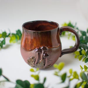 May include: A ceramic mug with a glossy brown glazed upper section and a matte, textured lower section. The base features sculpted, raised details of three mushrooms and fern leaves in a matching brown tone, complete with a curved side handle.