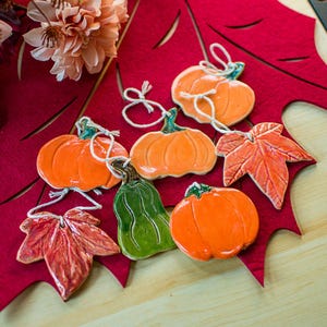 May include: A collection of ceramic ornaments featuring three orange pumpkins, one green gourd, and two textured autumn leaves. Each piece has a glossy finish and includes a white string loop for hanging, arranged together as a set of seasonal decorations.