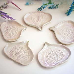 Peut inclure: Cinq plats en céramique faits à la main, en forme d'ail, de couleur blanc cassé, avec des surfaces texturées. Les plats sont disposés en cercle, mettant en valeur leur design et leur savoir-faire uniques. Les bords ont une teinte rose subtile.