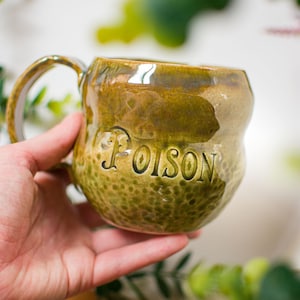 May include: A handmade ceramic mug with a glossy olive green glaze and a textured, dimpled surface. The word POISON is embossed in capital letters on the front. The mug features a curved handle and a rounded, organic shape with a smooth rim.