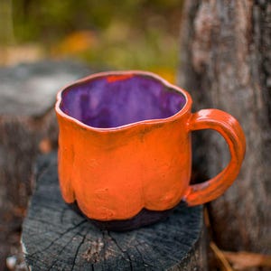Peut inclure: Une tasse en céramique faite à la main, avec un extérieur orange et un intérieur violet. La tasse présente une surface texturée et une base marron foncé. La poignée arrondie et la tasse posée sur du bois suggèrent un thème rustique ou automnal.