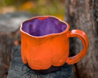 May include: A handmade ceramic mug with an orange exterior and a purple interior. The mug has a textured surface and a dark brown base. The rounded handle and the mug's placement on a wooden surface suggest a rustic or autumnal theme.