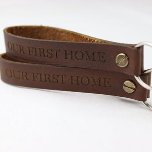 Puede incluir: Llavero de cuero marrón con dos llaveros y el texto "OUR FIRST HOME" grabado en cada lado.