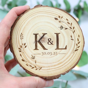 Può includere: Una fetta di legno con un bordo di corteccia naturale, che presenta un design inciso a laser con le iniziali "K&L" e la data "30.03.25" circondate da una ghirlanda floreale.