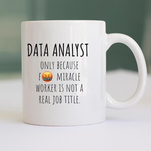 May include: White ceramic mug with the text "DATA ANALYST" in black, followed by "ONLY BECAUSE" and "MIRACLE WORKER IS NOT A REAL JOB TITLE." An angry face emoji is between the words "BECAUSE" and "MIRACLE". The mug has a curved handle.