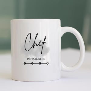 May include: White ceramic mug with the words "Chef In Progress" in black script. The mug features a progress bar with three filled circles and one empty circle, suggesting a work-in-progress theme. The mug has a curved handle.