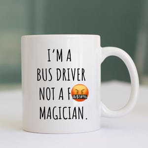 May include: White ceramic coffee mug with black text that reads "I'm a bus driver not a f&!&#% magician."  The mug has a black and yellow emoji with a red angry face and a black censor bar over the mouth.