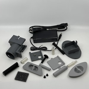 以下が含まれることがあります： 電源アダプター、さまざまなプラスチック部品、小さなフィルターなど、グレーと黒の電子部品の詰め合わせ。 部品は白い表面に配置されており、キットまたは関連アイテムのセットを示唆しています。