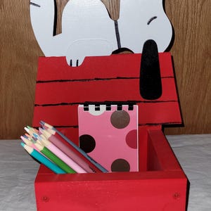 May include: A red wooden desk organizer shaped like Snoopy's doghouse. The white Snoopy figure is on top. Inside are colored pencils and a pink notepad with brown polka dots. A charming and functional desk accessory.